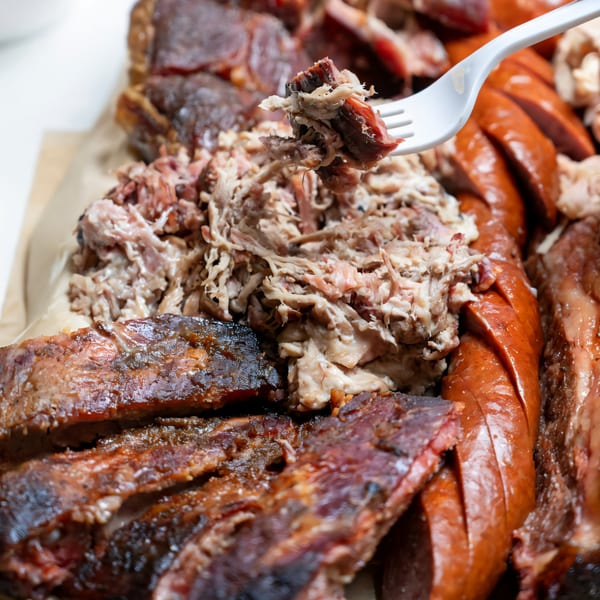 Close-up of a fork lifting pulled pork from a platter of Baby Back Blues BBQ ribs and sausage.