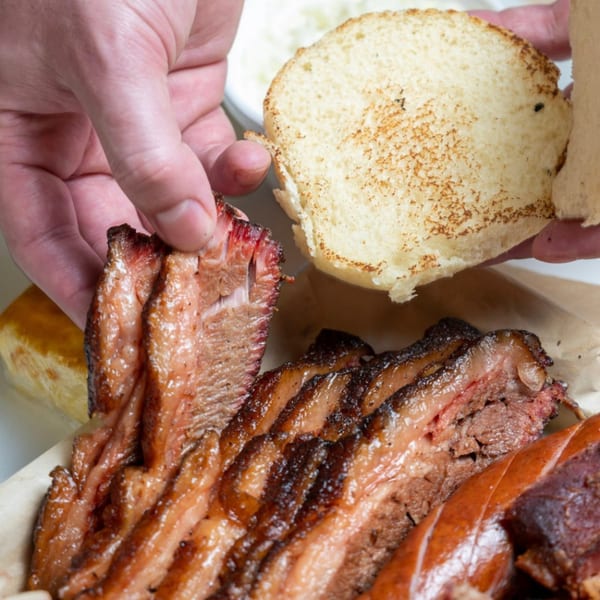 Hands preparing a Baby Back Blues BBQ brisket sandwich with toasted bun and sliced brisket.