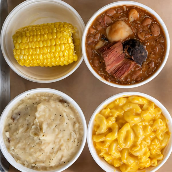 Four white bowls containing BBQ sides: corn on the cob, baked beans, mashed potatoes, and mac and cheese.